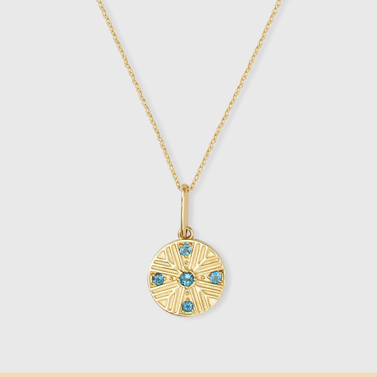 Runder Kettenanhänger aus 14k Echtgold im Art-Déco-Stil mit Sonnenstrahlen-Prägung und fünf blau funkelnden Topasen an einer feinen Goldkette vor hellem Hintergrund.
