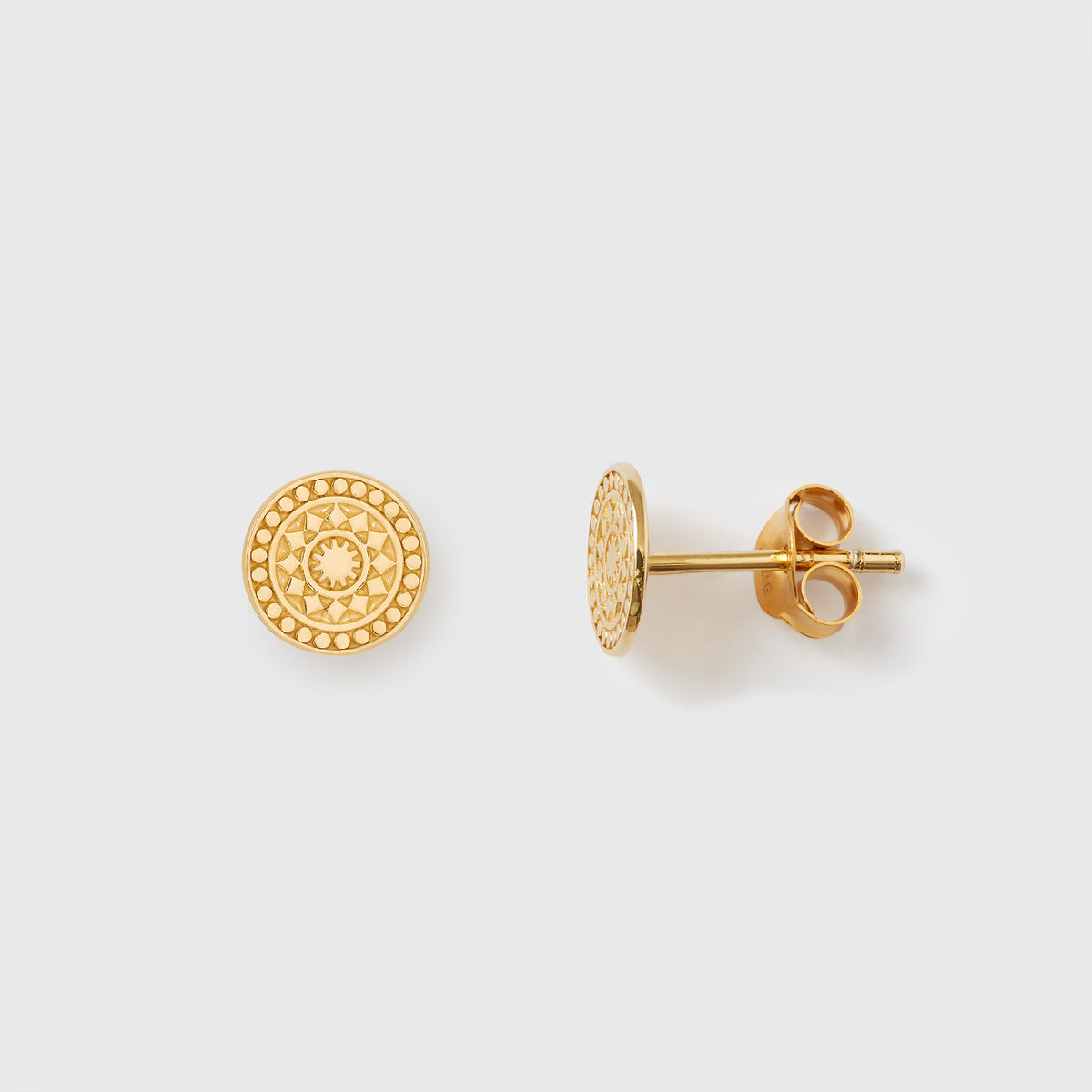 Nahaufnahme eines Paares goldener Ohrstecker mit feinem Ornament-Muster auf weißem Hintergrund. Der linke Ohrstecker zeigt die flache, kreisrunde Vorderseite mit detaillierter Ornament-Prägung, der rechte liegt seitlich und zeigt den Steckerstift sowie den Verschluss.